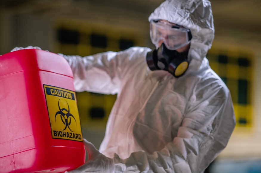 Person im Schutzanzug hält Kanister mit Aufdruck Biohazard vor sich.